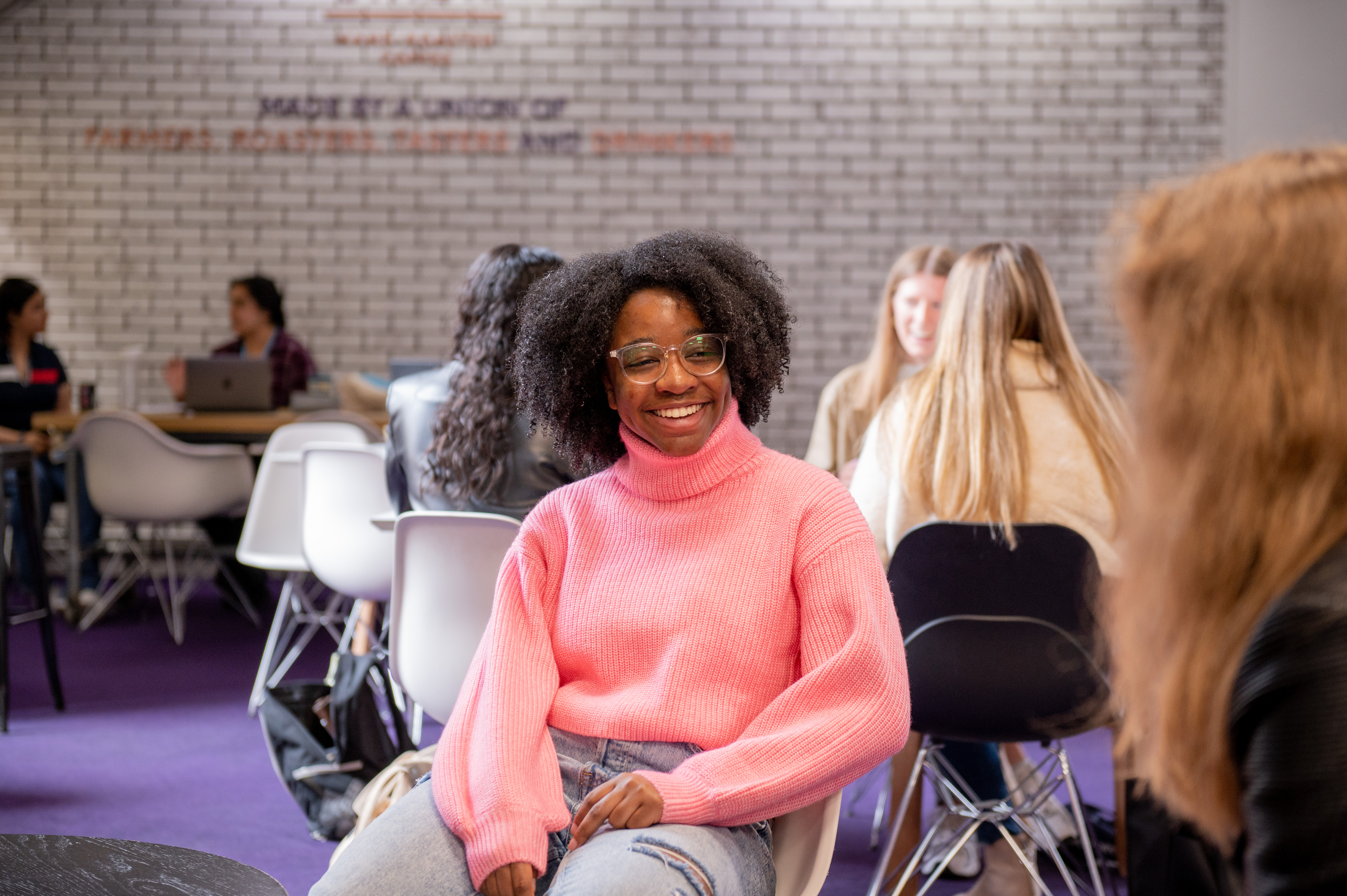 Students sit around in several small groups having discussions. At the centre a student in a pink jumper is facing the camera and smiling