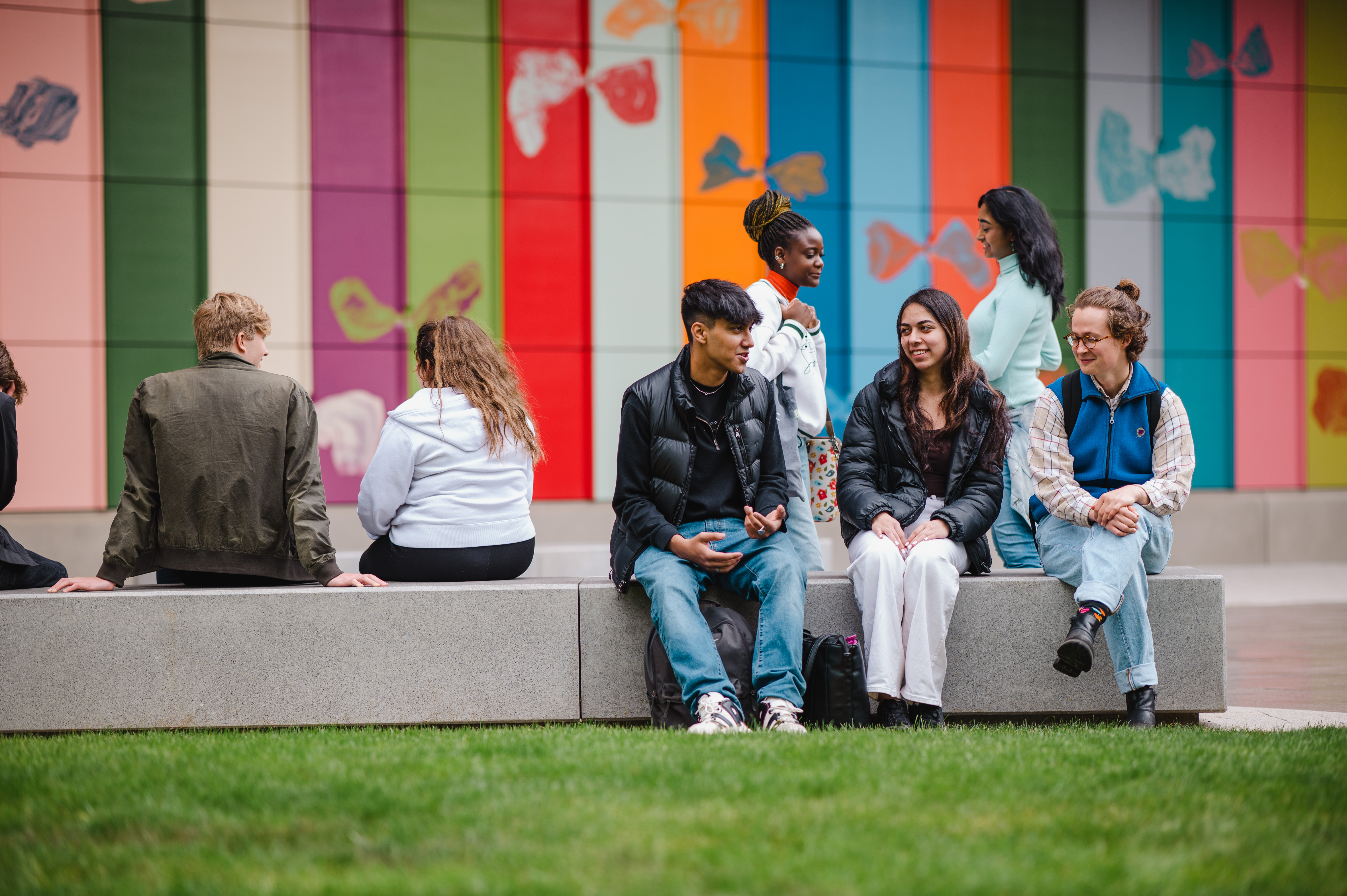 Several students sit outside the FAB, a multicoloured artwork is in the background