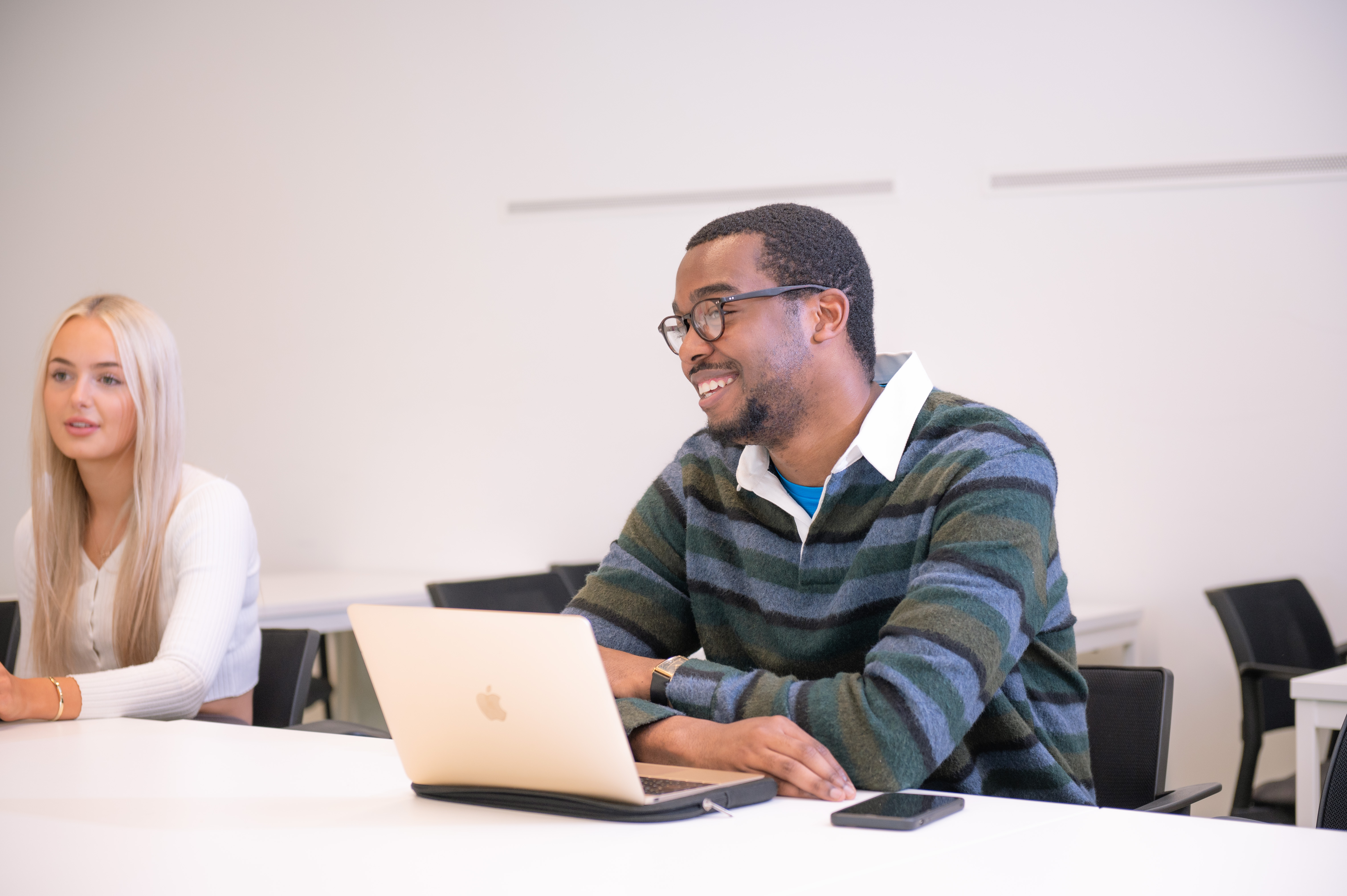 A Philosphy student smiles as they talk through he work they are doing on their laptop while a second student with long blonde hair listens carefully