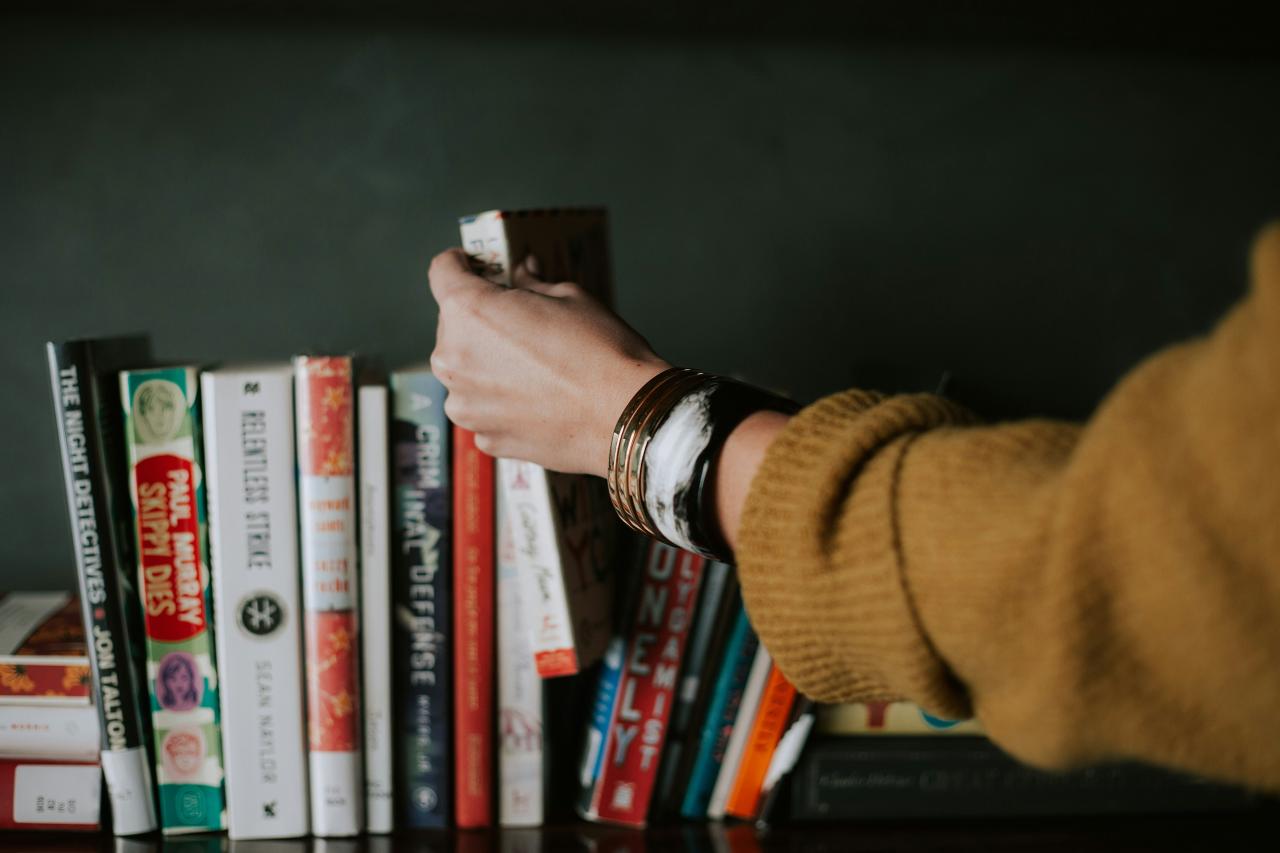 A hand reaches out to take a book out of a row of books along a shelf. 