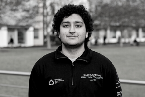 A black and white photo of a man smiling at the camera. He has thick, dark, curly hair and is wearing a dark, polo tshirt.