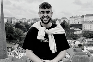 A black and white photo of a man smiling at the camera. He has short, dark hair, a beard and moustache, white skin and dark eyebrows. He has a pale jumper tied around his shoulders and is wearing a black tshirt.