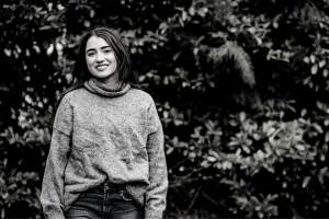 A black and white of a young, white woman wearing a polo neck jumper and jeans, standing in front of a hedge. The woman has dark hair and is smiling.