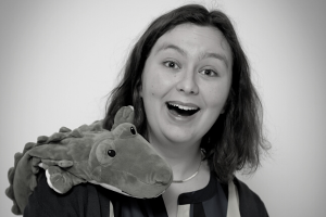 A black and white photo of a smiling woman with dark hair and white skin. She looks incredibly happy, and also has a soft toy on her shoulder - a fluffy crocodile.