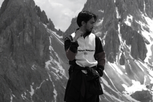 A black and white photo of a man standing in front of a mountain range. He has dark skin, is wearing climbing gear and is looking off to the right hand side, with one hand by his face.