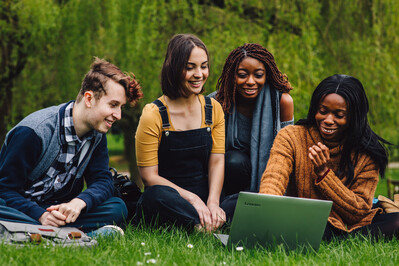Four students are sat outside on the grass with two laptops, all four are looking at one laptop screen and laughing together.
