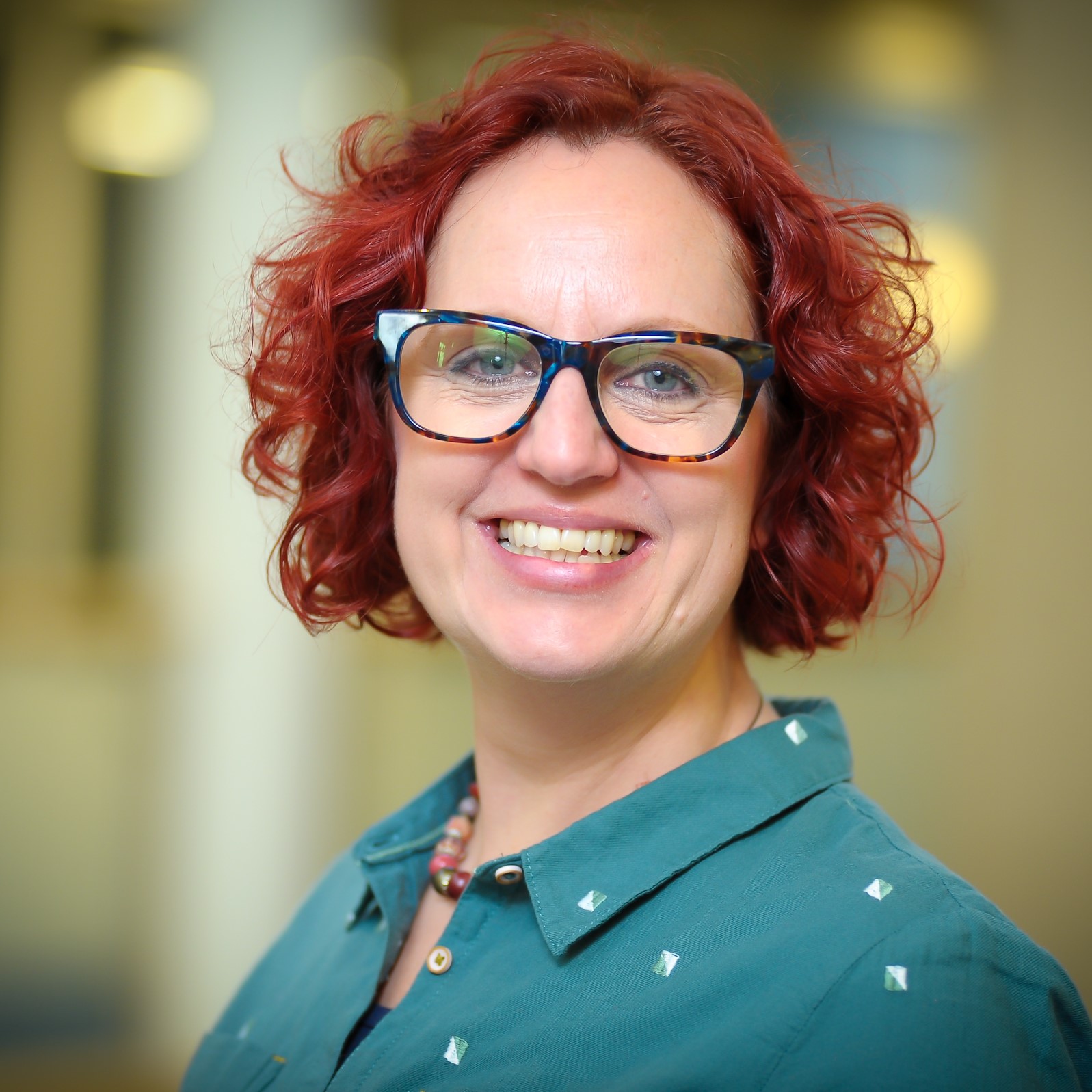 Image of a woman with short curly red hair in black rimmed glasses on a blurred background.
