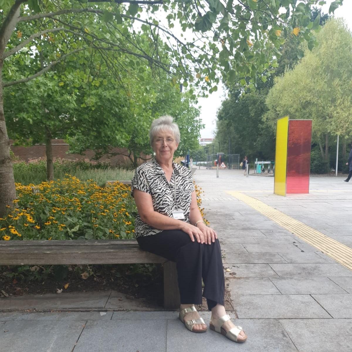 Image of a woman with short hair and glasses sitting on a bench with greenery and trees in the background.
