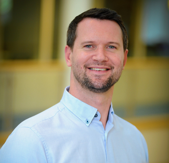 Image of the head and shoulders of a man in a blue collared shirt on a blurred background
