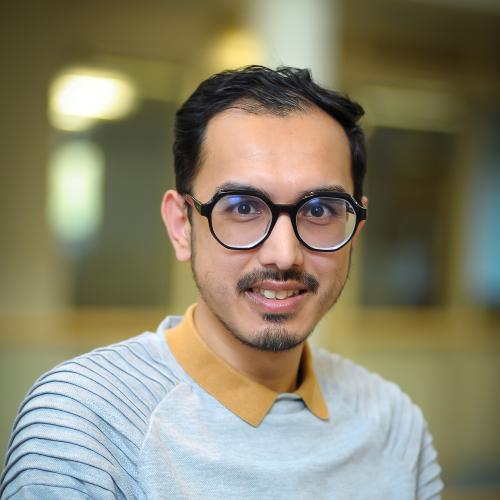 Image of a man in a light blue top with a mustard yellow collar on a blurred background