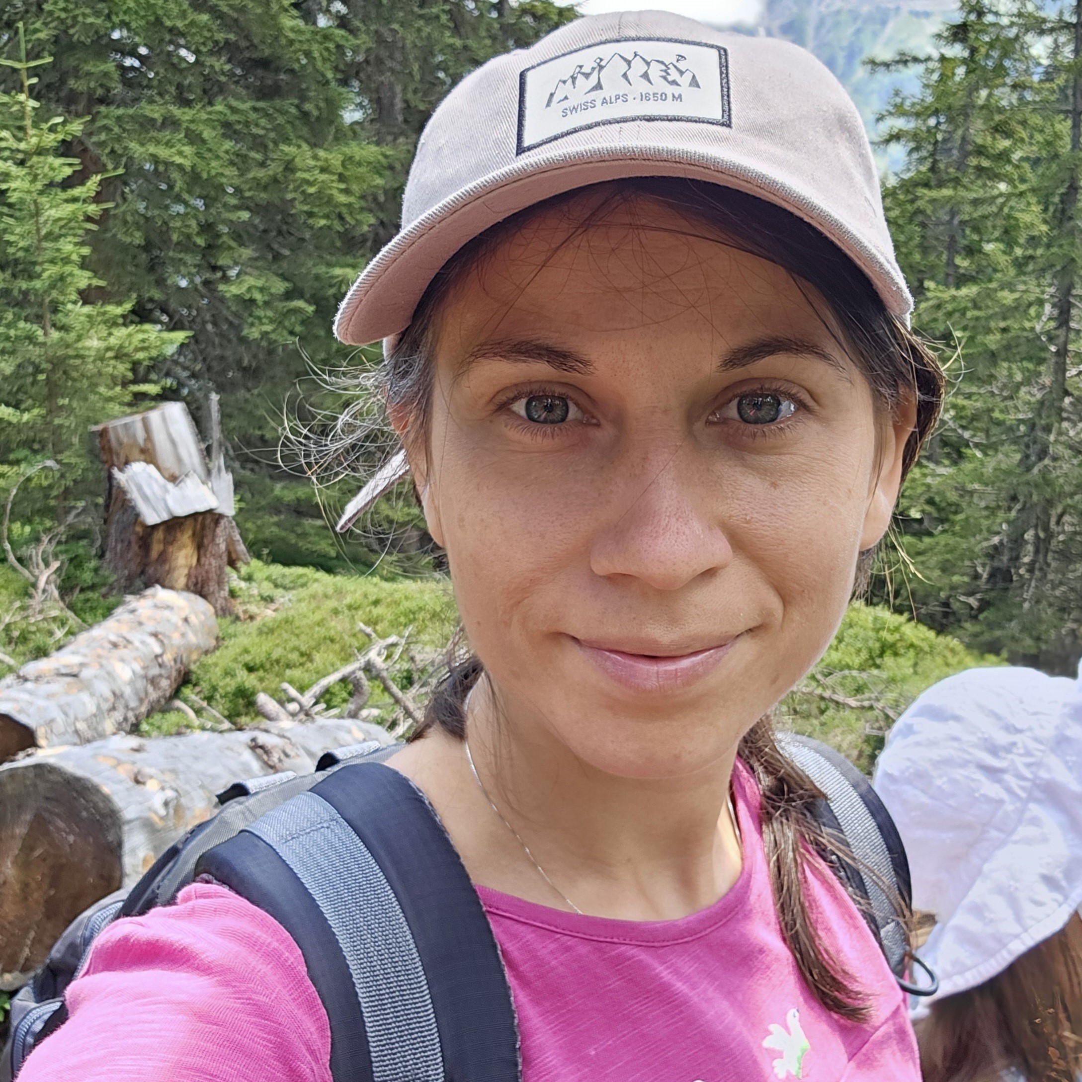 Image of a woman in a bright pink top wearing a backpack, with long brown hair in a ponytail over her shoulder and a white sun cap covernig her head. In the background is a forest.