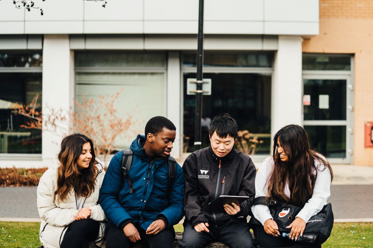 A group of 4 students sitting on a bench on a piece of grass outside the front of building.