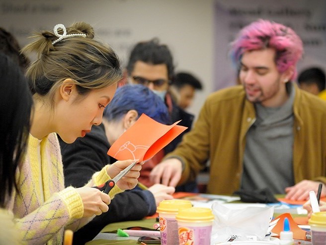 student cutting out a piece of orange card whilst another student with ink hair decorates next to them.