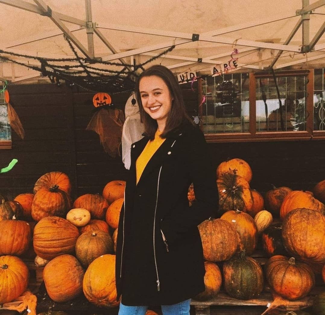 Image of Michelle Watons wearing a yellow jumper, black long coat and jeans. In the background stands a large mound of pumpkins.
