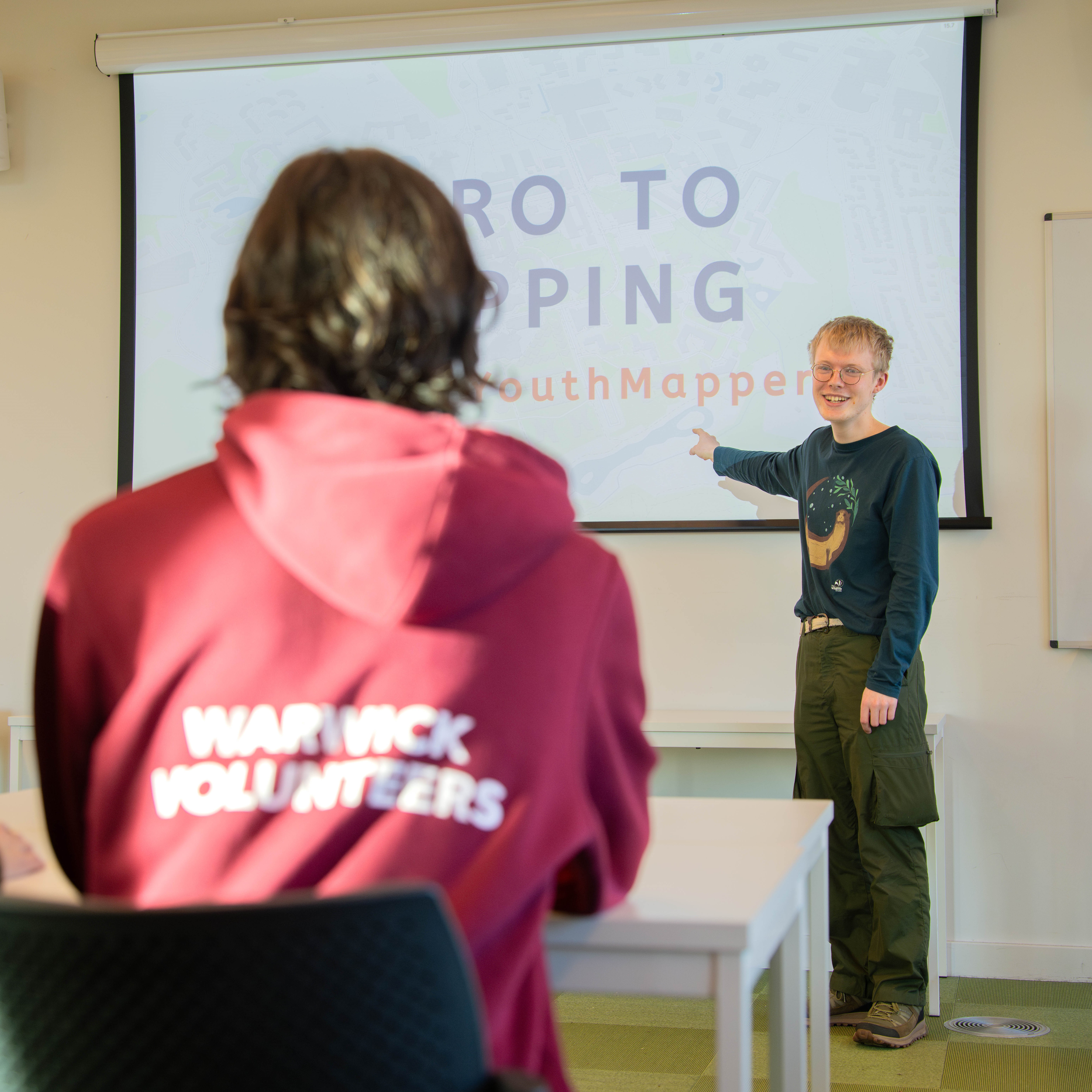 Image of a person standing at the front of a room pointing to a screen, with someone in a burgundy Warwick Volunteers hoodie sitting in the foreground