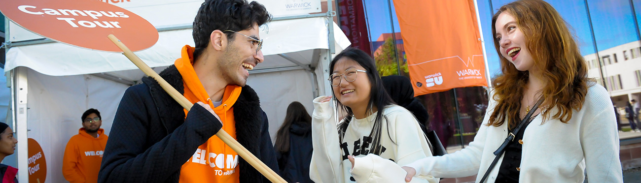 A student Welcome Helper holds a lollipop emblazoned with "Campus Tours", as they chat with to new students, laughing.
