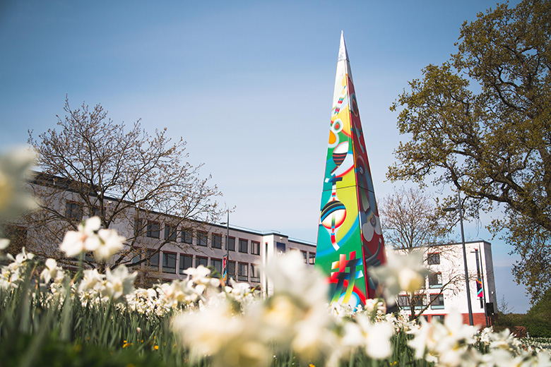 Sculpture 'Needle of Knowledge Obelisk' in front of the Ramphal Building, blue skies above, daffodils in the foreground