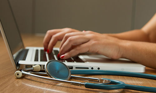 A hand types on a laptop next to a stethoscope