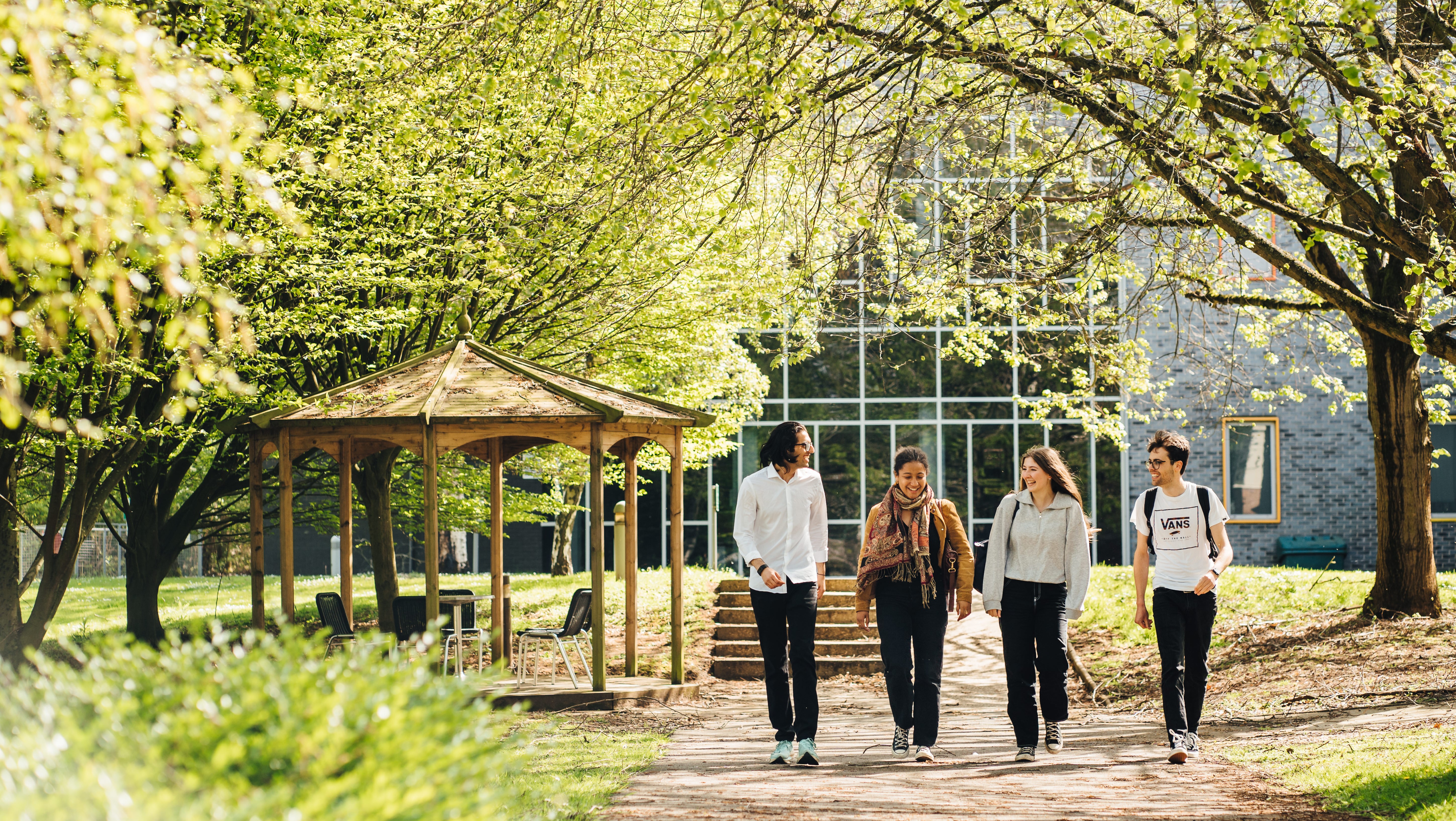 Image of 4 students walking along a garden path, with a wooden gazebo on their left