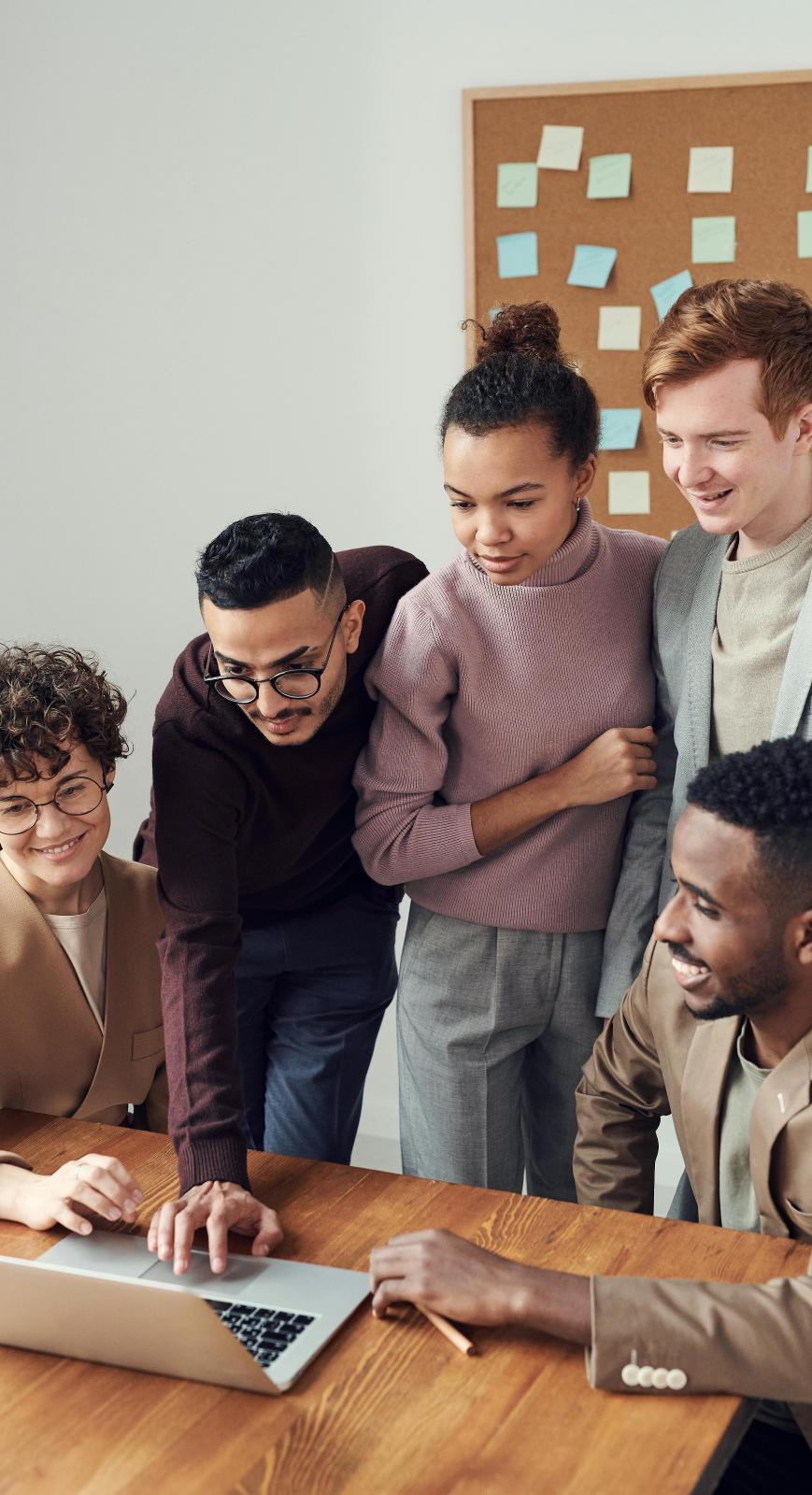Group of 5 people crowded around a laptop, a corkboard is visible in the background