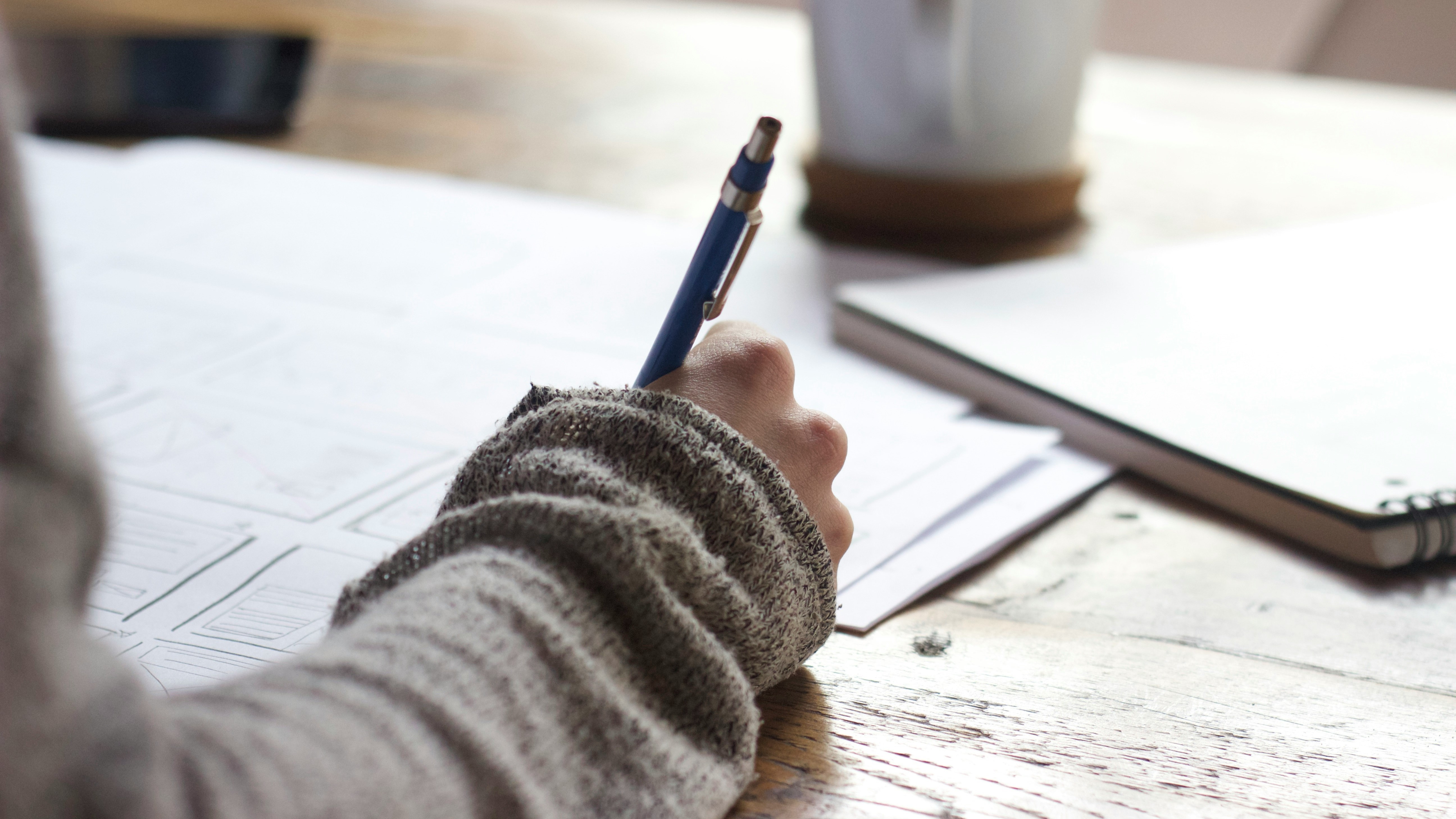 Person writing on a piece of paper with notebook and mug in background