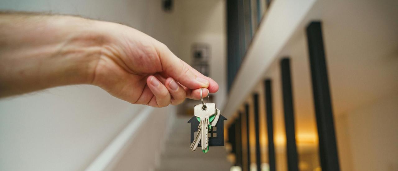 Hand holding a set of house keys, with a house shaped key chain, blurred in the background is a set of stairs leading up