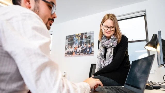 Two people in business wear sat around a laptop having a discussion.