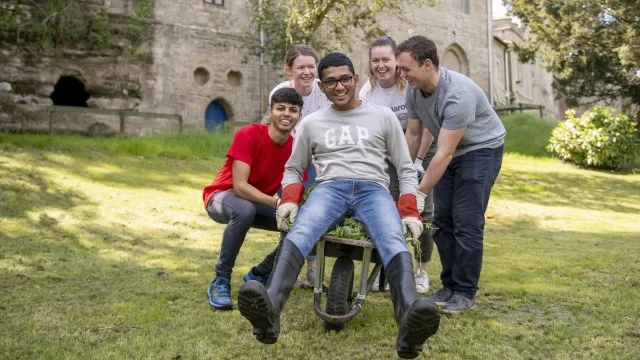 One student is sitting in a wheelbarrow laughing whilst four others are gathered around pushing him and laughing.