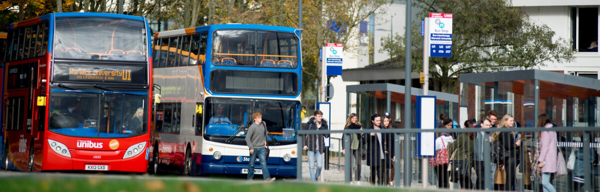 Students standing a a bus stop, with two buses parked on the left. 