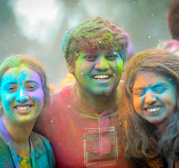 Three students smile at the camera, snow floats through the air and they grin with their faces coated with a rainbow of coloured powder.