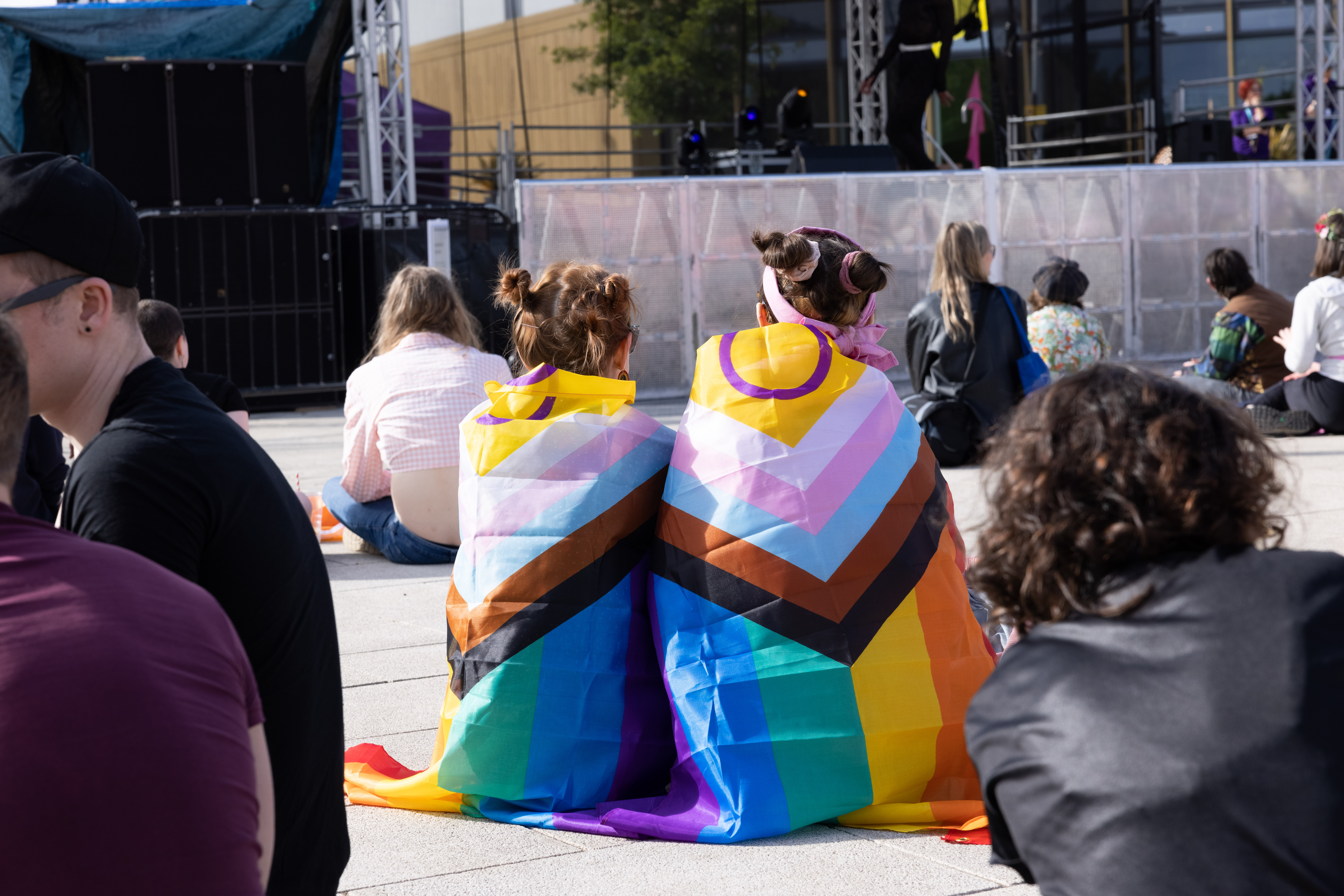 two people sit on the ground together wrapped in the Pride flag. 
