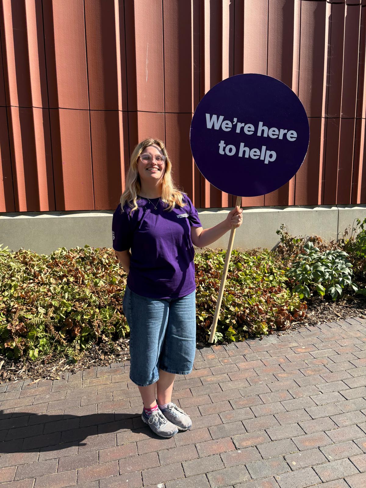 welcome helper in purple uniform holding a here to help sign