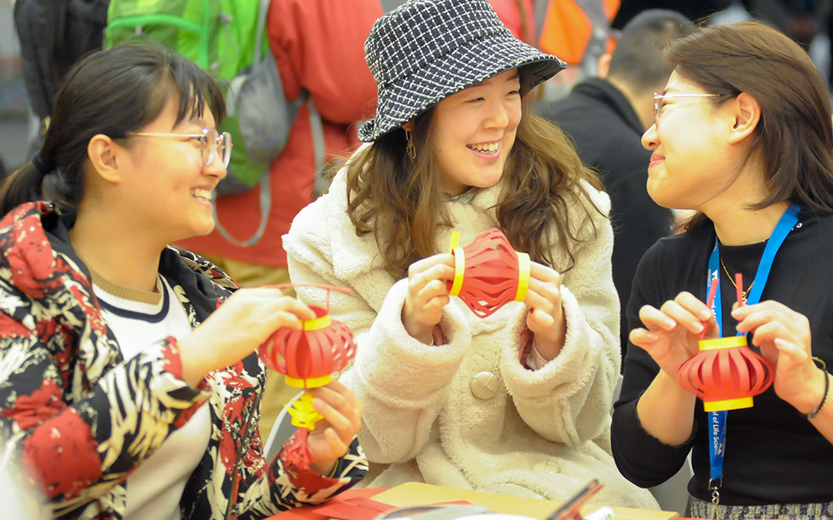 A group of students look at each other and smile as they make paper lanterns at a workshop