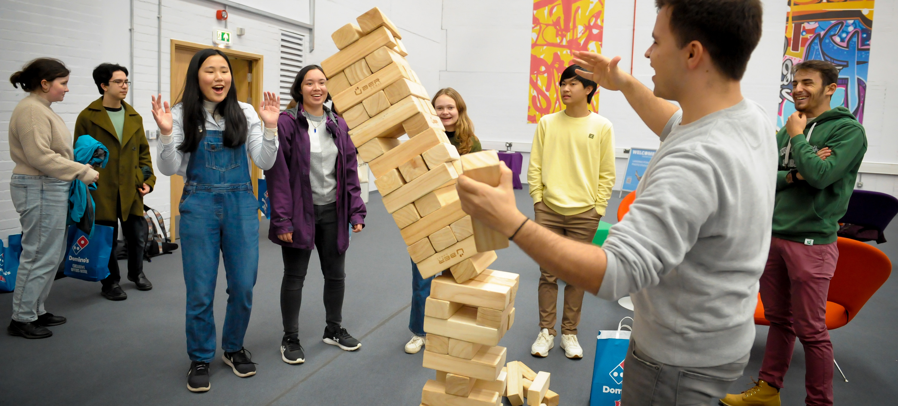  A group of students playing giant Jenga, the blocks are falling towards a female student who raises her arms in surprise.