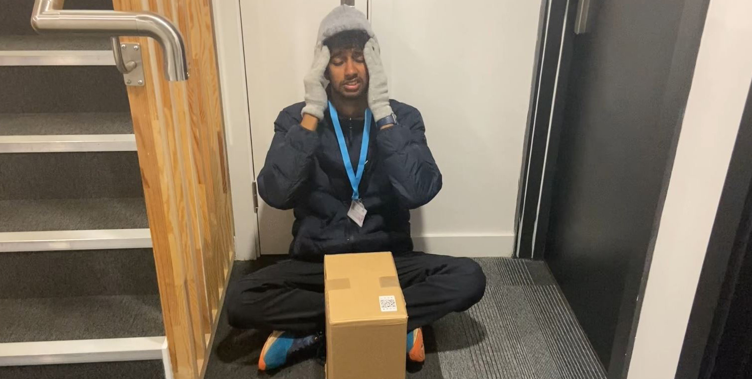 A delivery driver sits on the floor, head in his hands with a box by his feet.
