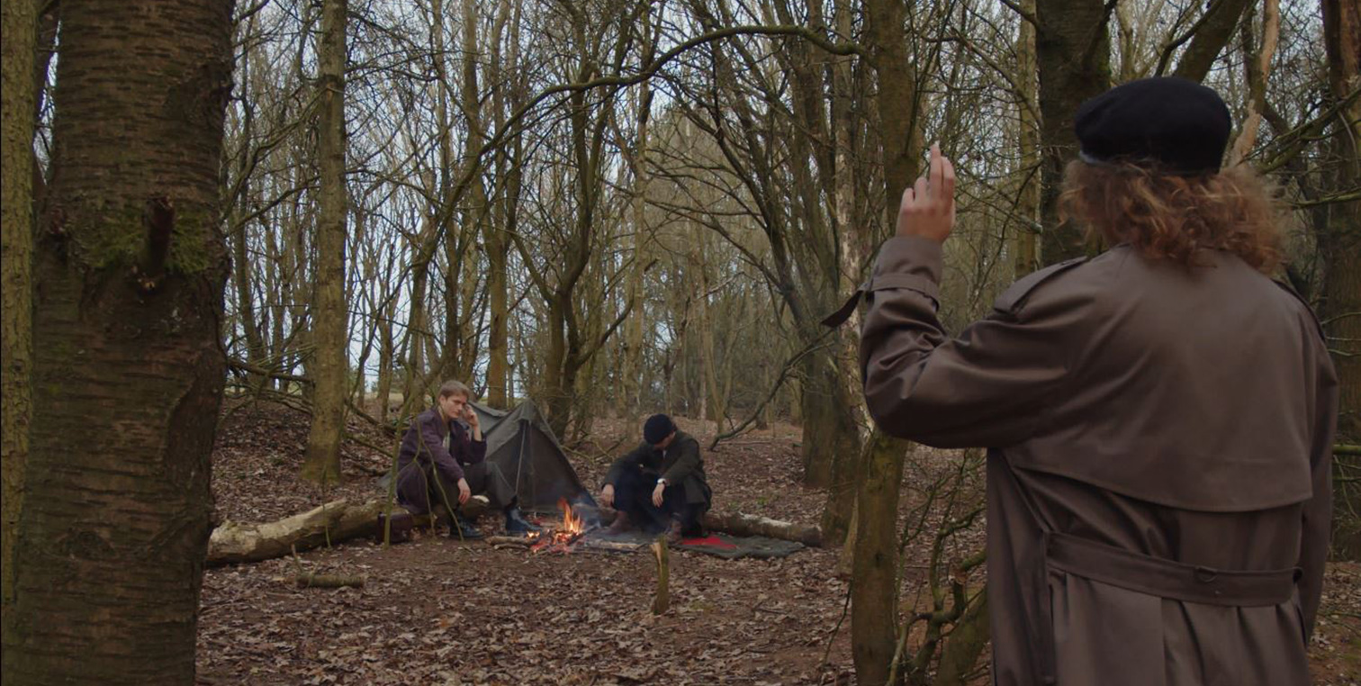 Two men sit glumly at a campfire in the woods, a woman approaches and raises her hand.
