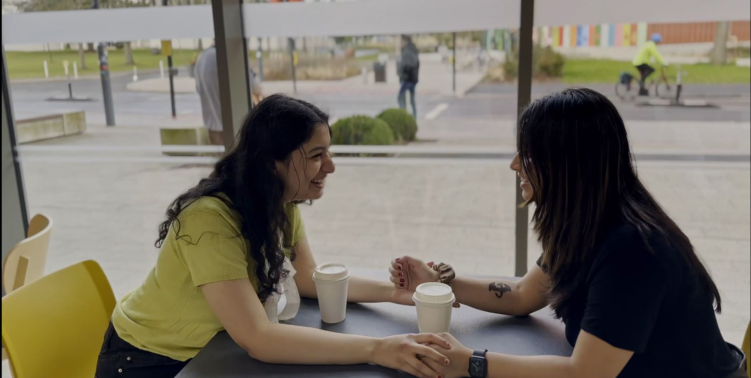 Two women are chatting animatedly at a coffee date, clasping hands across the table.