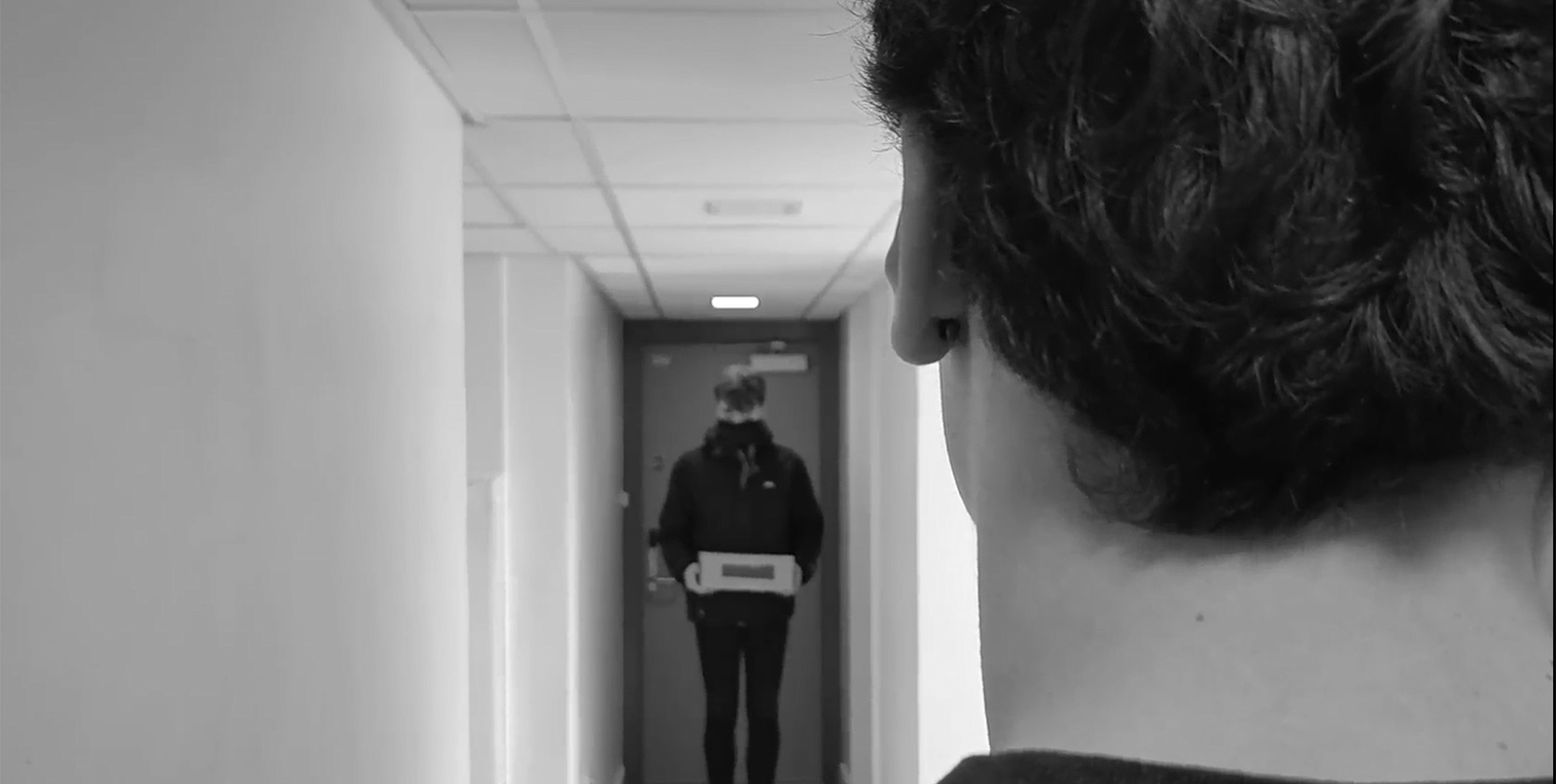 A man stands ominously in a hallway, holding a box.