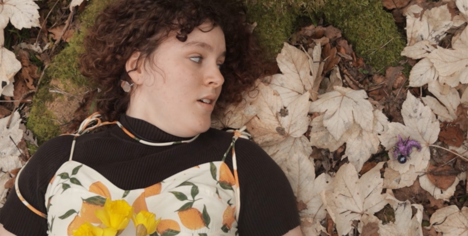 A woman lies on a bed of leaves, looking at a toy dragon.