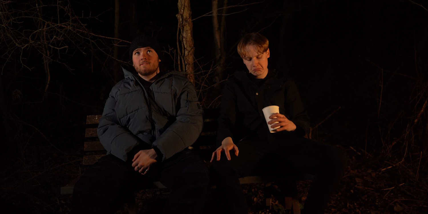 Two men sit on a bench in the talk, one looks to the sky whilst the other inspects his coffee cup.