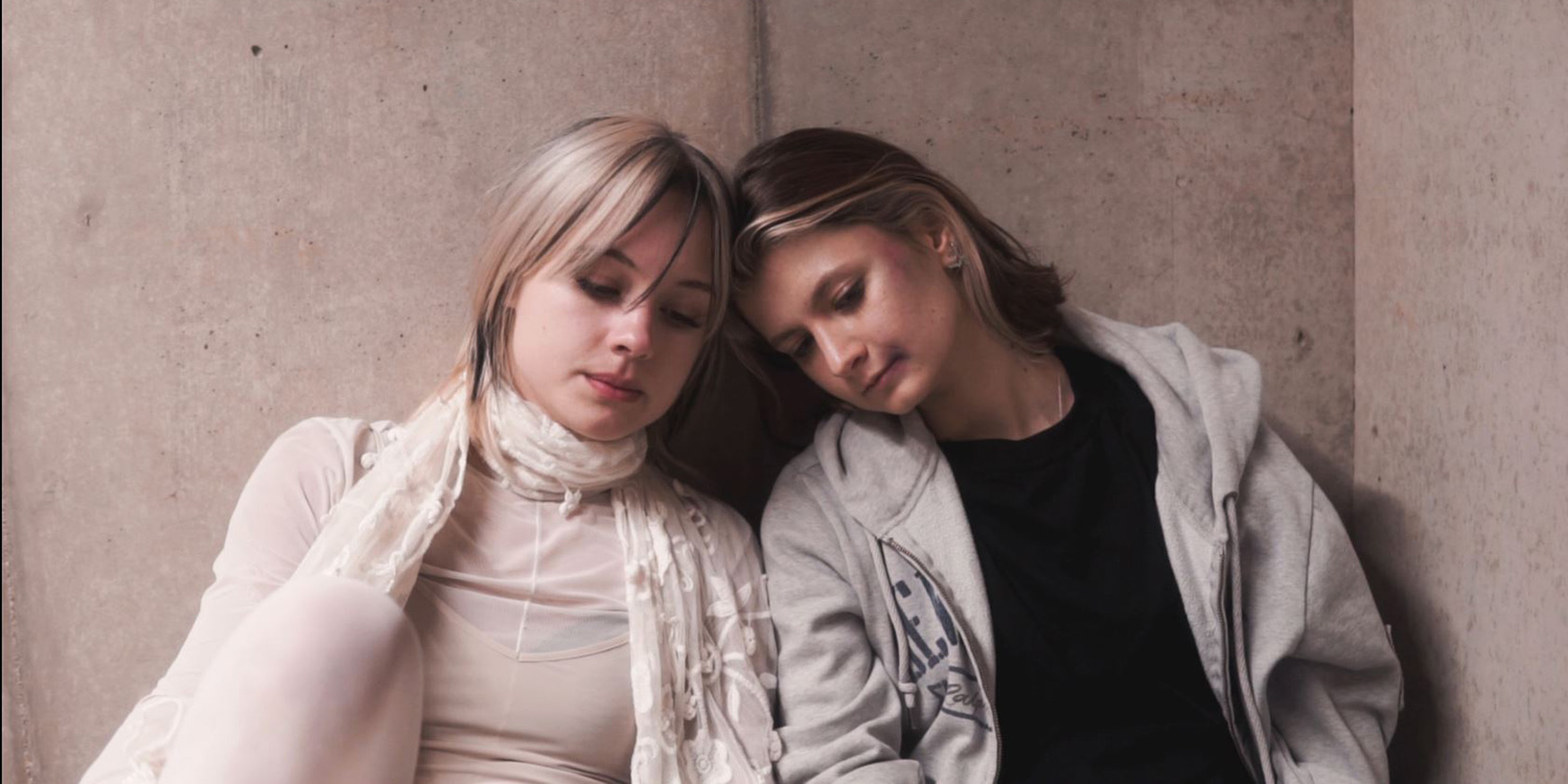 Two women sit leaning against a concrete wall, their heads touching, looking defeated.