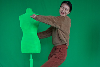 A student dances with a manequin in front of a green screen, smiling at the camera.