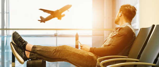 A student reclines with their feet on top of their suitcase at the airport, a plane seen taking off in the background.