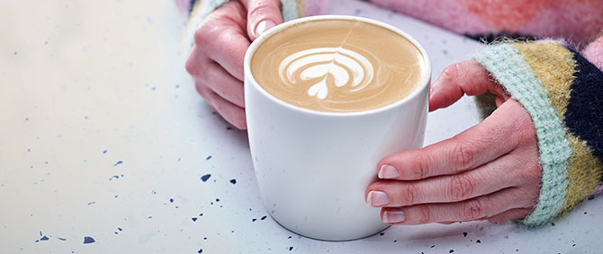 A hand in a brightly coloured wooly jumper holds a cup of coffee, with a leaf shape poured into the milk.
