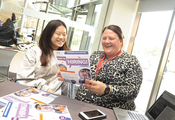 A member of staff laughs with a new student as they look a leaflet, which reads "We're Hiring! Widening Participation"