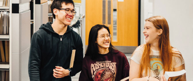 Three students chat and laugh carrying books through a corridor