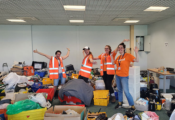 A group of students in high vis jackets pose with their hands in the air surrounded by materials donated for their swap shop