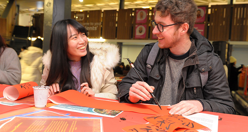 Two students smile at each other as one teaches the other calligraphy