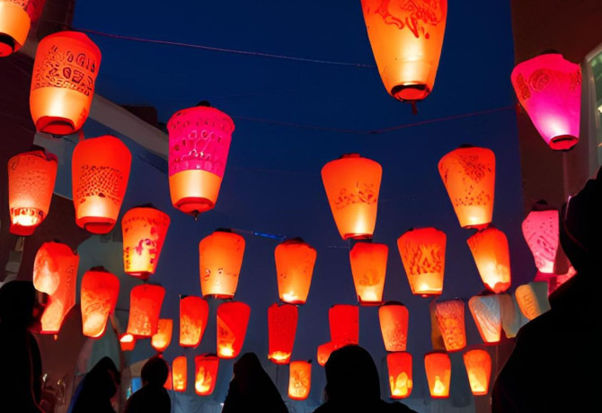 A silhouetted crowd looks at red paper lanterns hanging between buildings against a dark blue night sky.