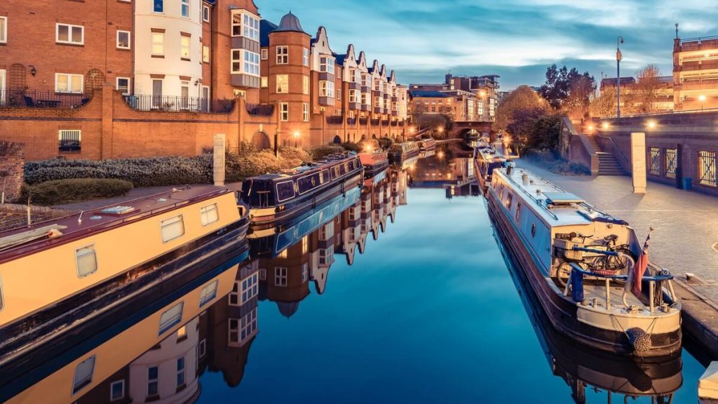 The picture is of a canal at night, the waterfront is lit up, making the water reflect the night sky and appear like glass. Canal boats are moored on either side of the canal. The image gives off a feeling of rest and tranquillity as no people are pictured